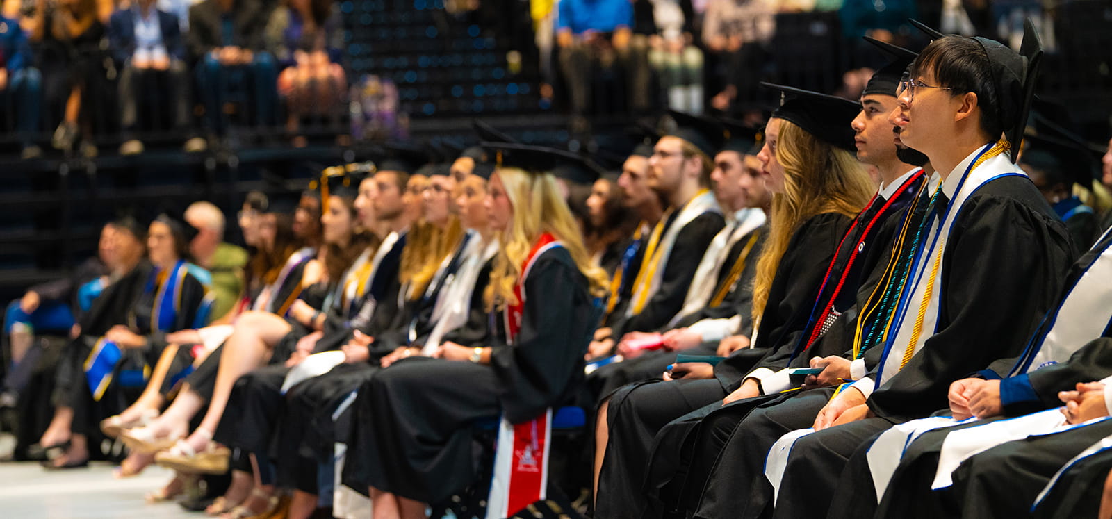 students sit at a graduation ceremony wearing their caps and gowns