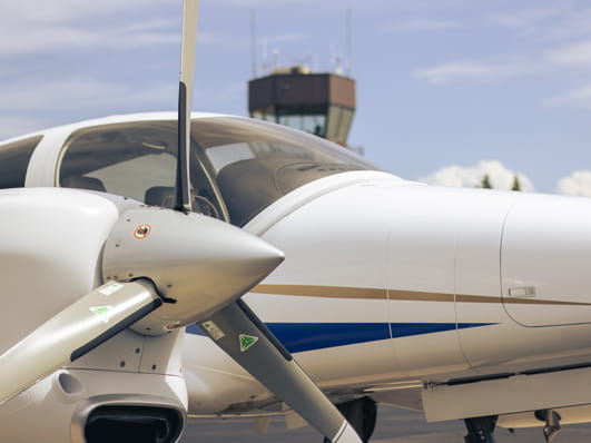 Close-up of a small white aircraft with its propeller in the foreground and an air traffic control tower visible in the background.
