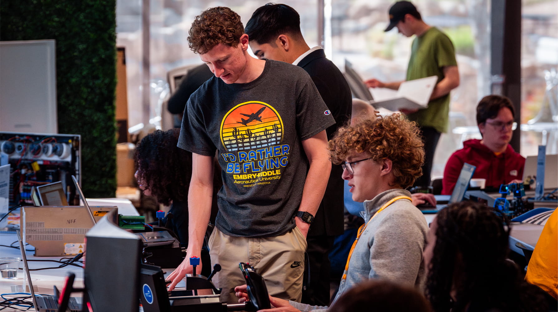 A group of people gathered around computers and equipment at an indoor event, with several attendees working and talking in the background.