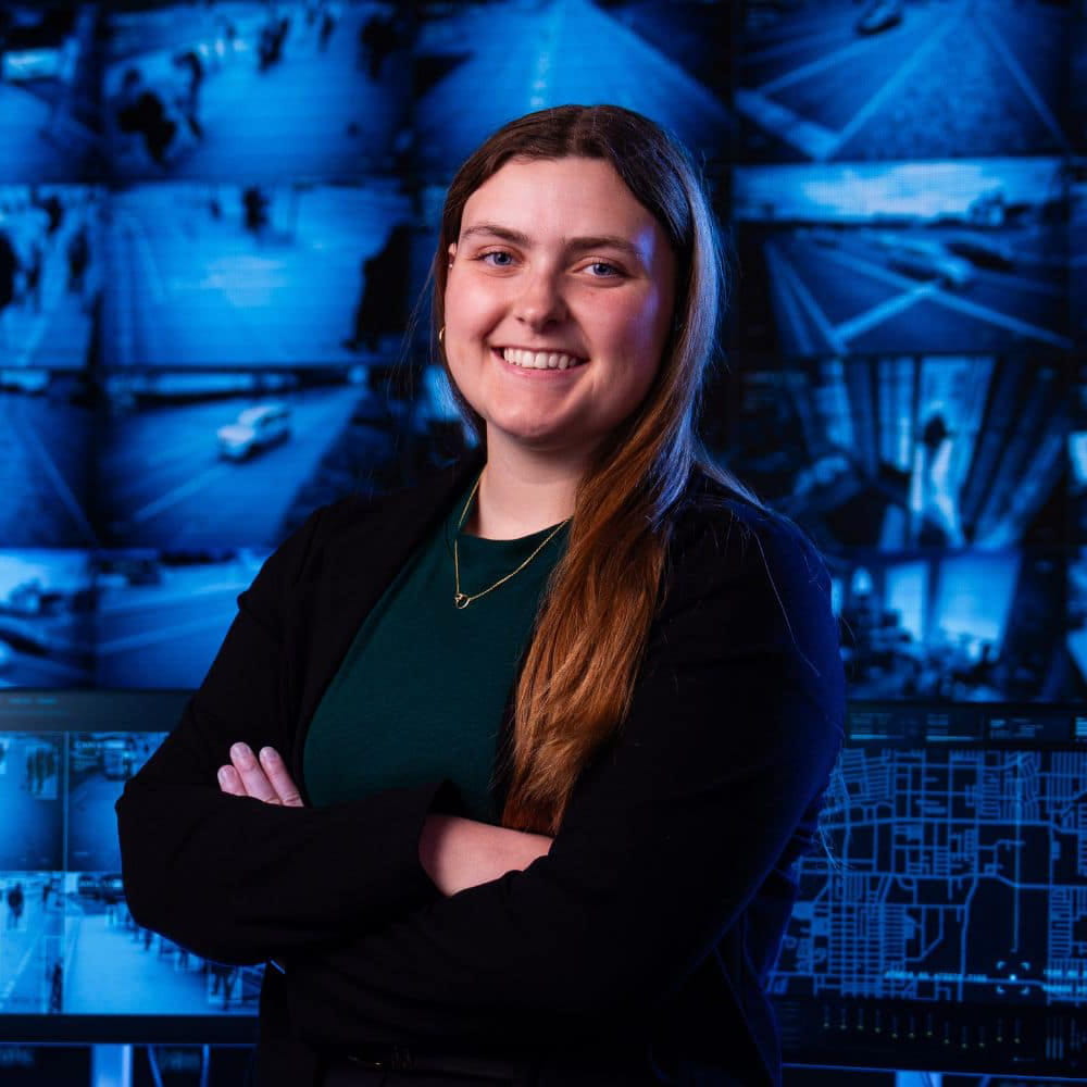 A student stands with arms folded in front of a wall of blue surveillance camera monitors.