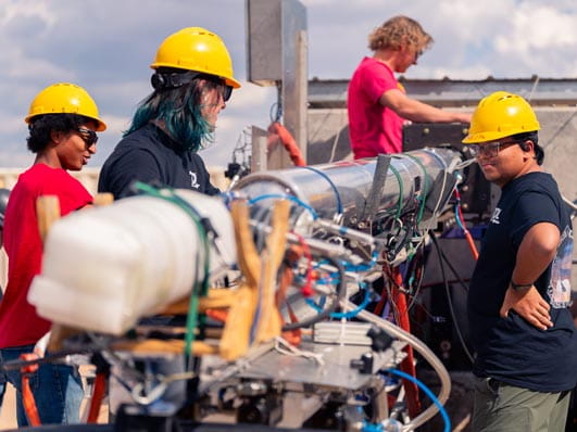 Group of people wearing hard hats working on a large, complex piece of machinery outdoors.