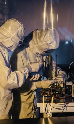 Two people in full protective suits working with scientific equipment in a lab setting.