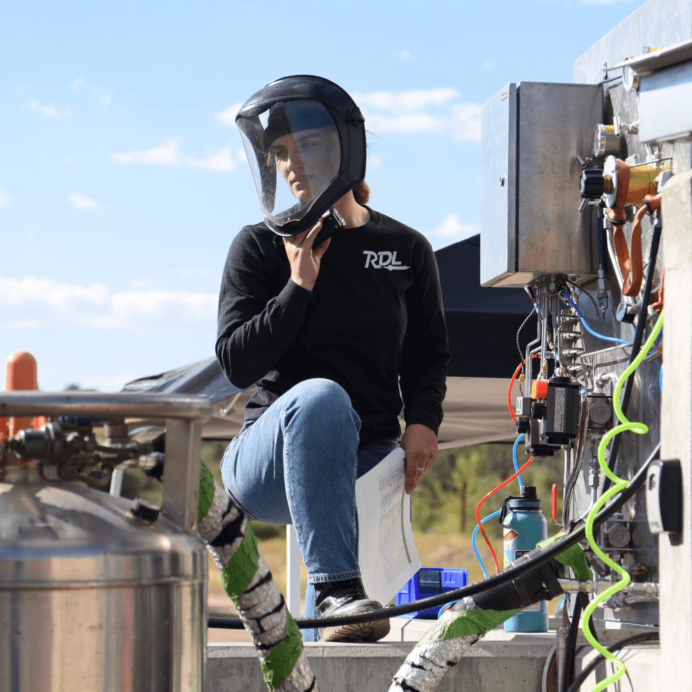 Olivia Wood wears a welding helmet and stands amid welding equipment