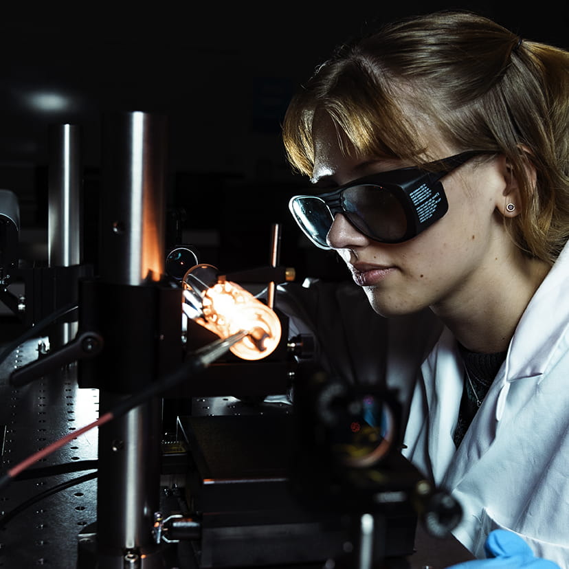 Embry-Riddle student in a lab coat and safety glasses works in a lab examining a lightbulb