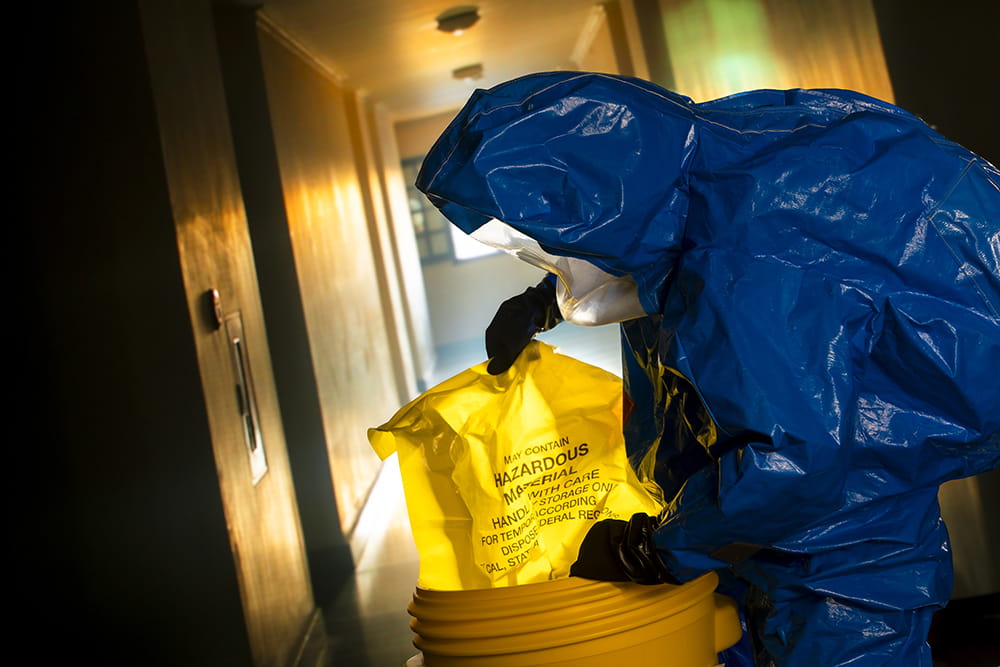 a person in a hazmat outfit places a bag of hazardous materials in a bucket for containment