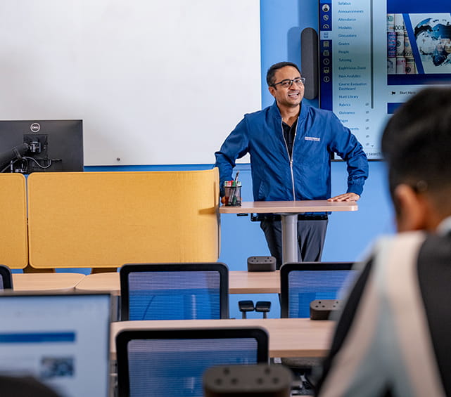 A person standing at the front of a classroom giving a presentation, while students with laptops listen and take notes.