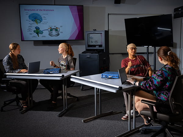 A small group of people sitting around tables in a classroom or meeting room, having a discussion with laptops open and a presentation displayed on a screen in the background.
