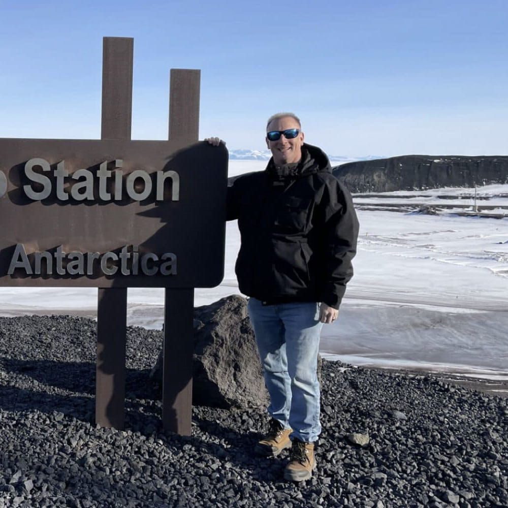 Marc Tunsall stands in front of a sign for a station in Antartica in front of an icy tundra