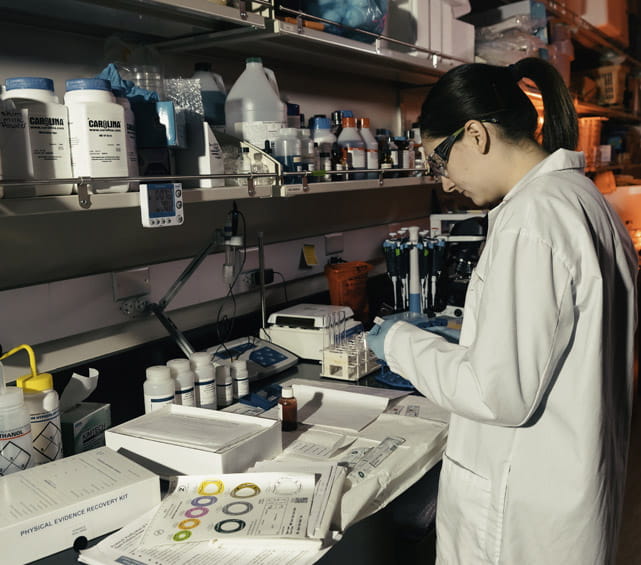 A scientist wearing a lab coat and safety glasses works at a laboratory bench surrounded by equipment, chemicals, and test tubes.