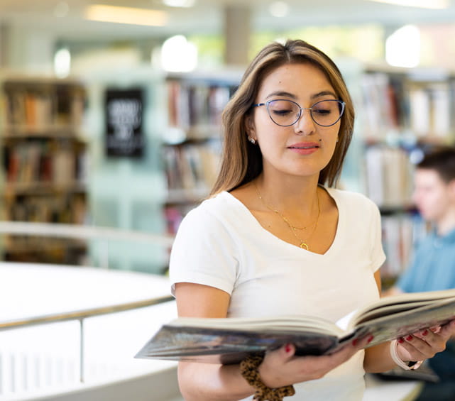 A person standing in a library while reading a book.