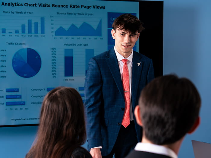 Embry-Riddle student in a business suit stands at the front of a room presenting analytics