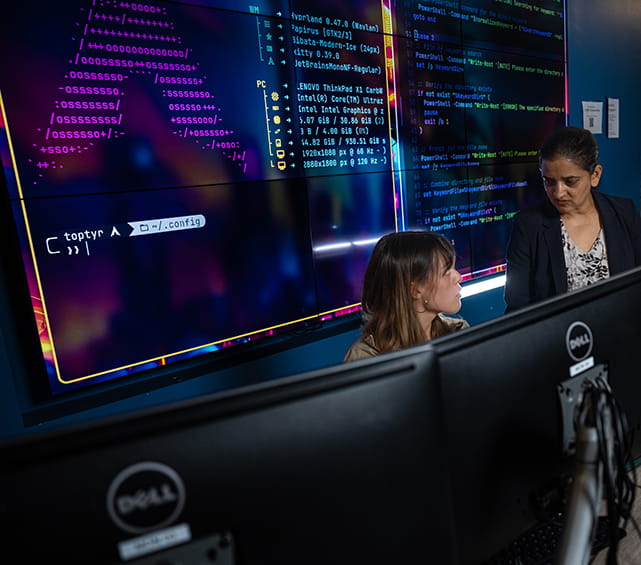 Embry-Riddle faculty member and student talk in the CyberLab with a screen of code behind them