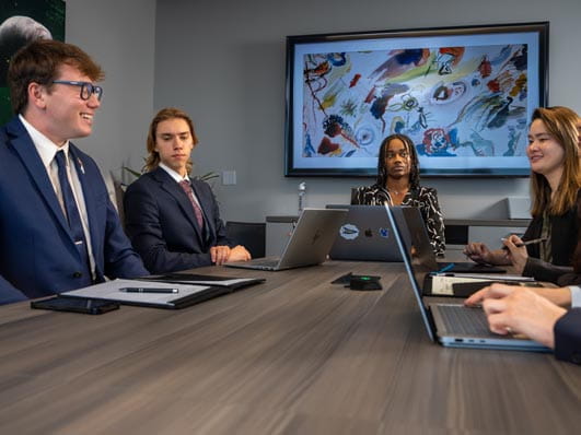 A group of five people sitting around a conference table in a meeting room, using laptops and talking.