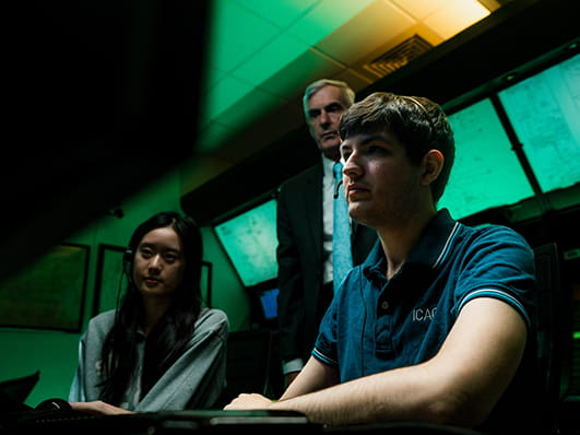 Two Embry-Riddle students sit in a lab with screens behind them as a professor looks on