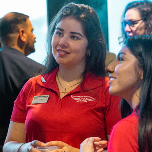 Two students in red Embry-Riddle polos talk with others at an indoor event.