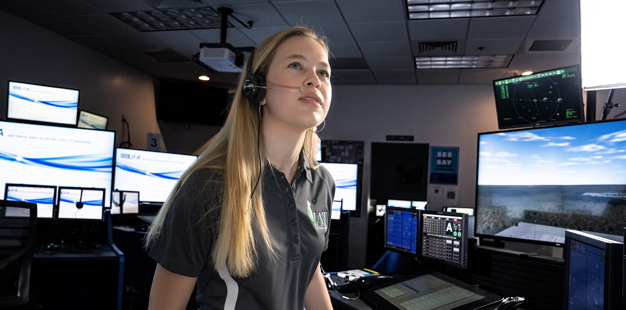 Woman wearing a headset operates controls in an aviation simulation lab surrounded by multiple monitors.
