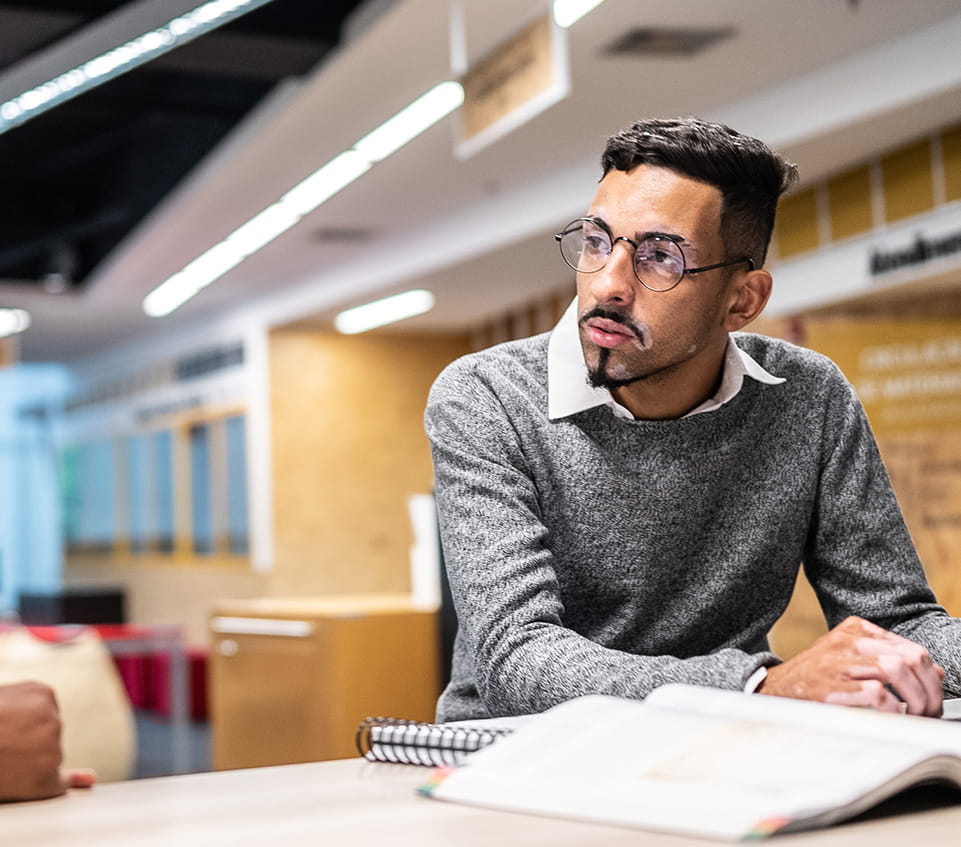 Student wearing glasses looks to the side while seated at a table with an open textbook and notebook in a campus study space.