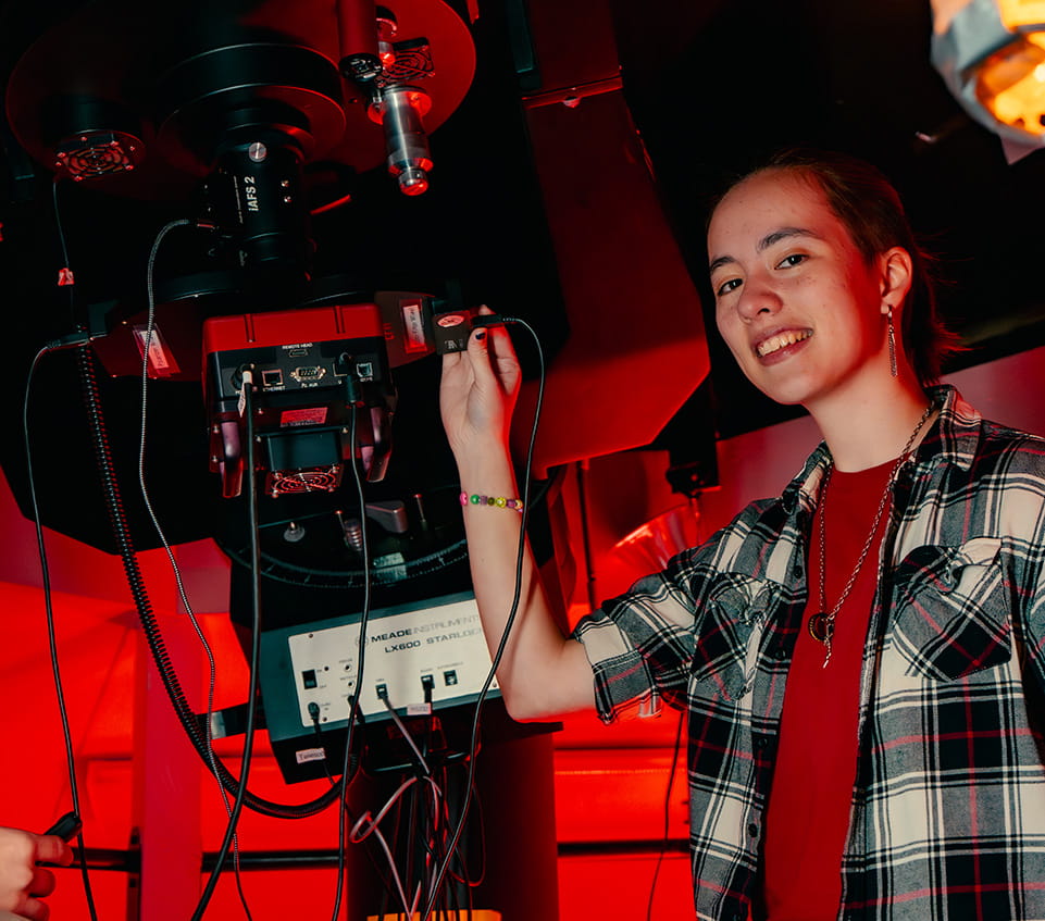 Student connects a cable to a Meade LX600 StarLock telescope control unit, adjusting imaging equipment beneath the mounted telescope in an observatory setting.