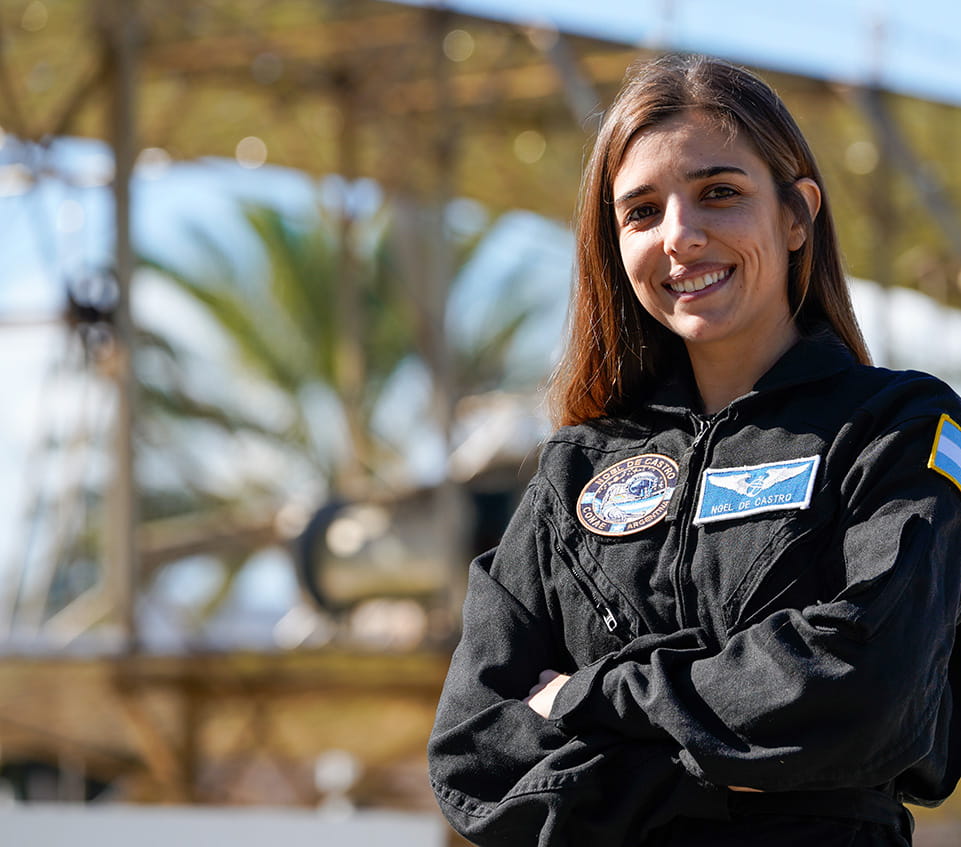 Woman in a flight suit with mission patches stands with arms crossed in front of the Wright brothers plane sculpture on the Daytona Beach Campus.