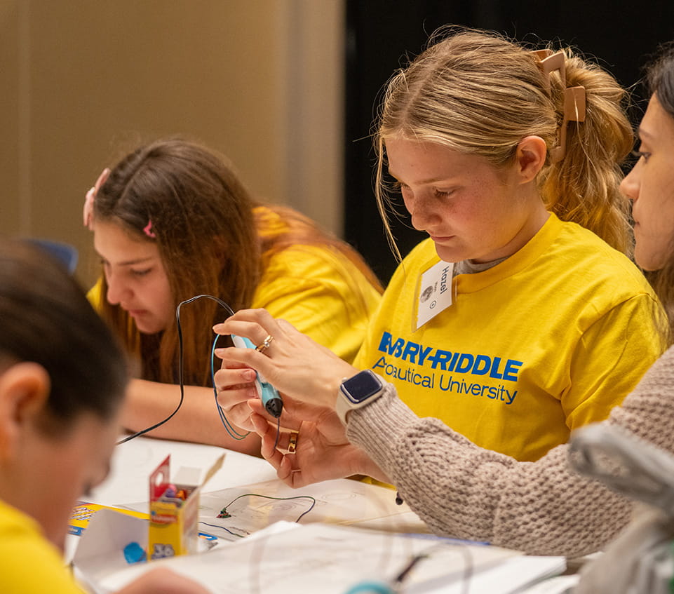 Students wearing Embry-Riddle t-shirts assemble a simple electronics project, connecting wires to a small motor or circuit on a table.