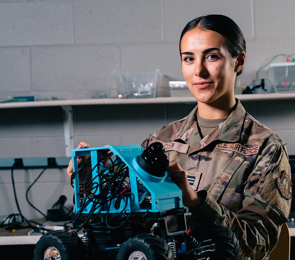 U.S. Air Force cadet holds a small robotic rover with exposed wiring and mounted camera module in a workshop setting.