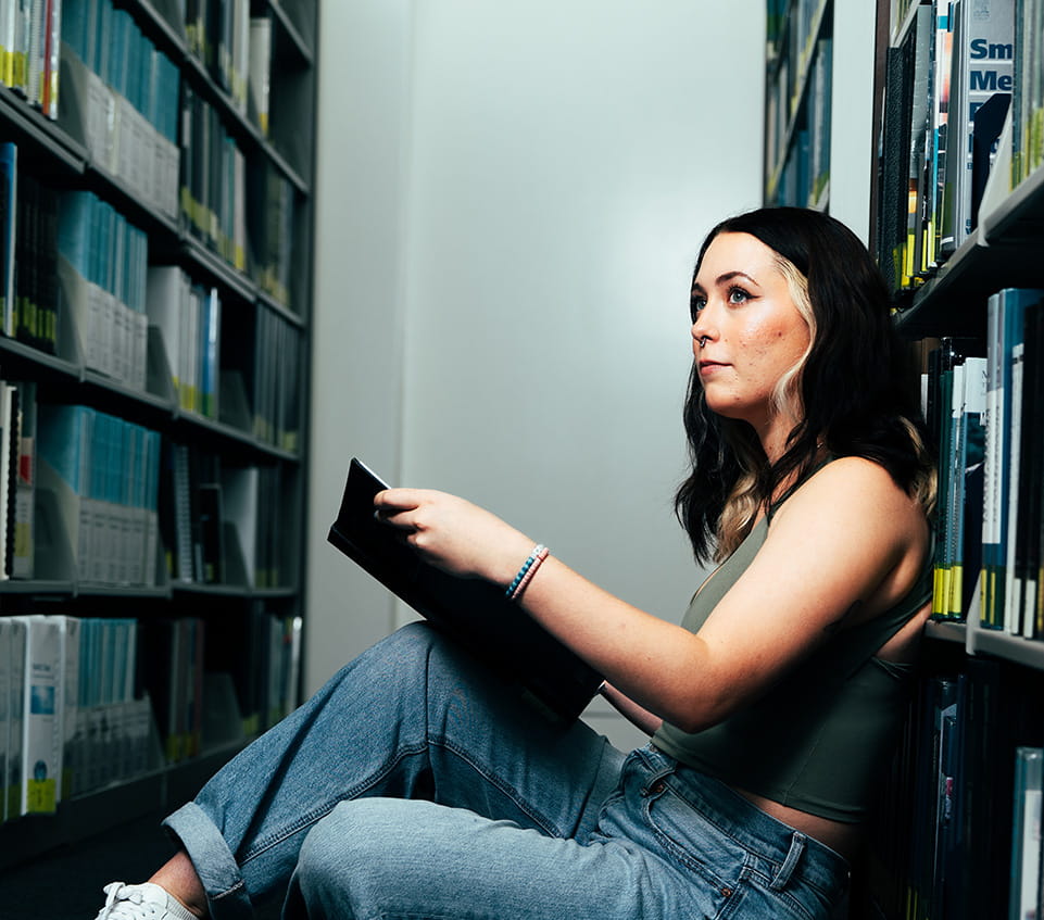Student sits on the floor between library shelves, looking up from a book.