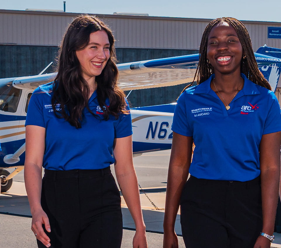 Two students in blue polo shirts walk and smile away from a single-engine training aircraft on the ramp.