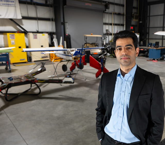 A person wearing a dark suit jacket and light blue dress shirt stands in an aircraft hangar, facing the camera.