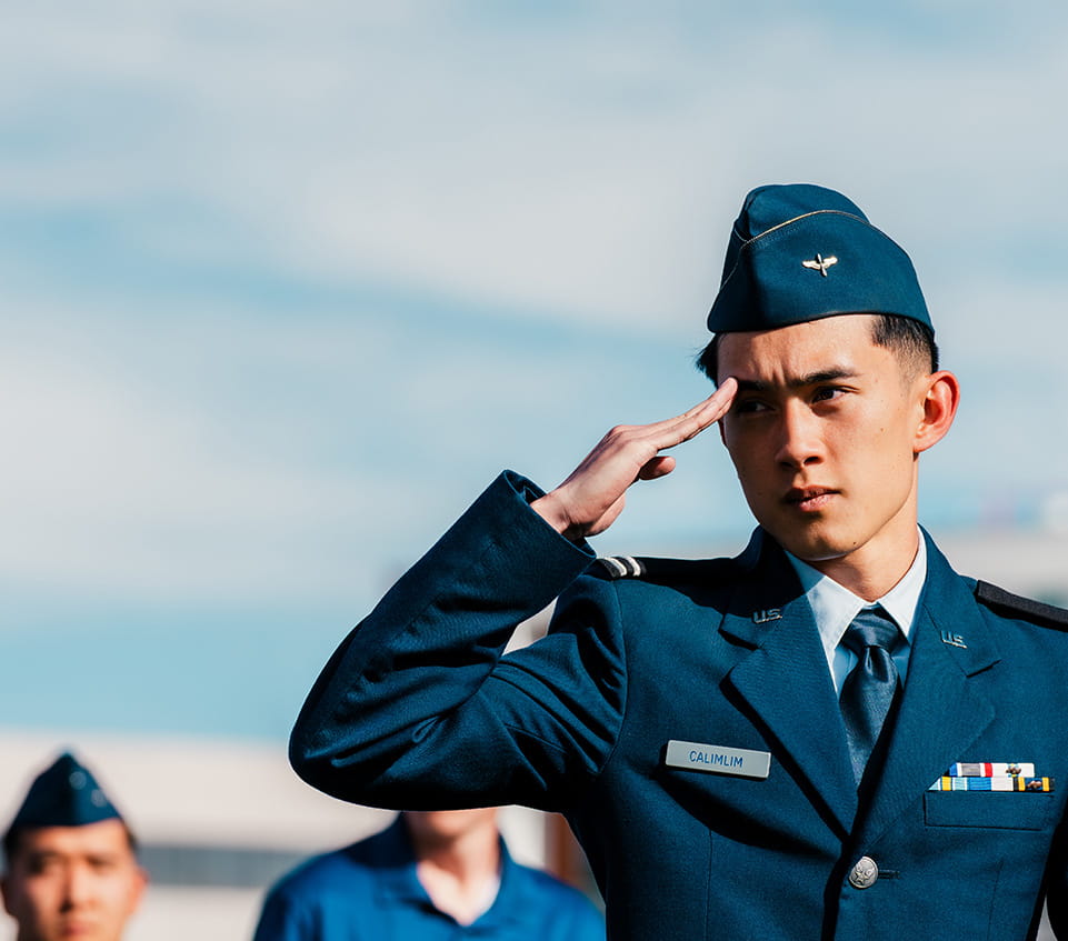 U.S. Air Force service dress uniform with ribbons and flight cap, young man saluting with right hand at brow, name tag visible on chest, other uniformed individuals blurred in background.