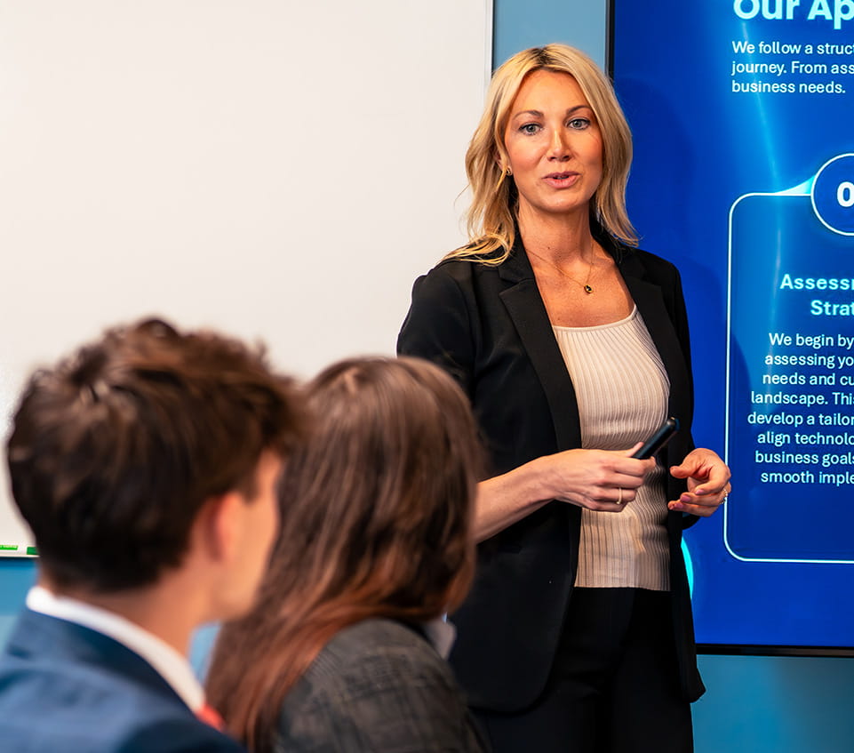 Faculty member leads a discussion beside a large display showing a process diagram and text, addressing seated students in a classroom.