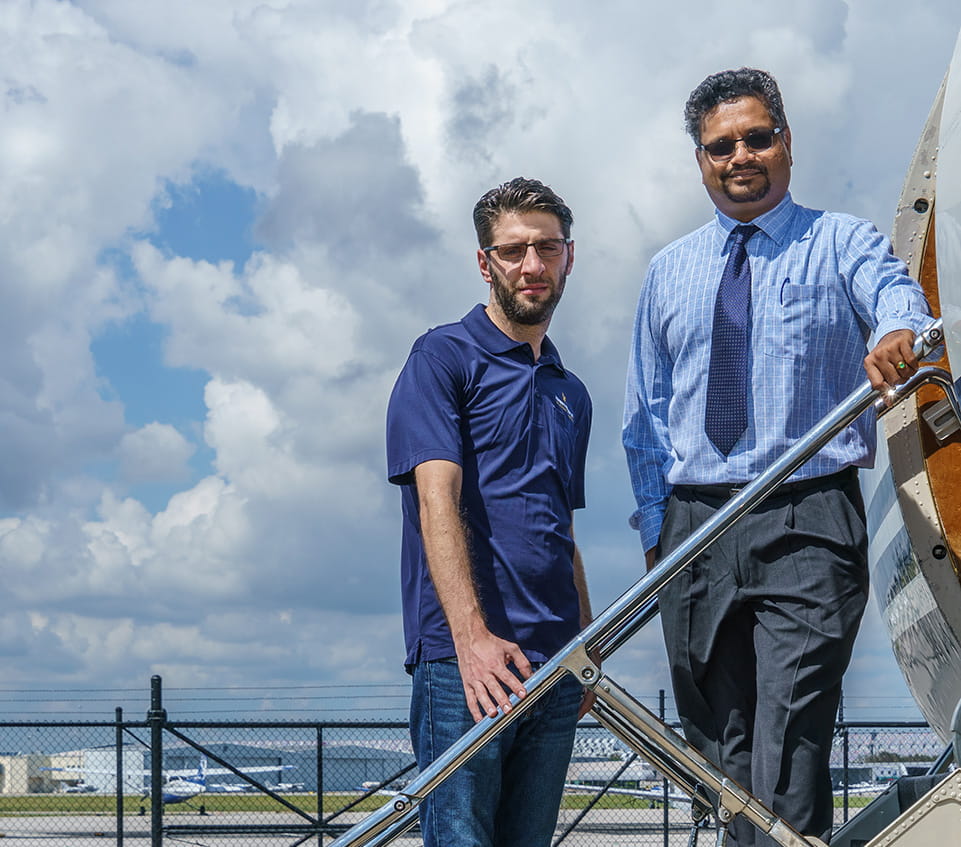 Two men stand on aircraft stairs beside a small airplane on the ramp, with airport fencing and parked aircraft under a cloudy sky.