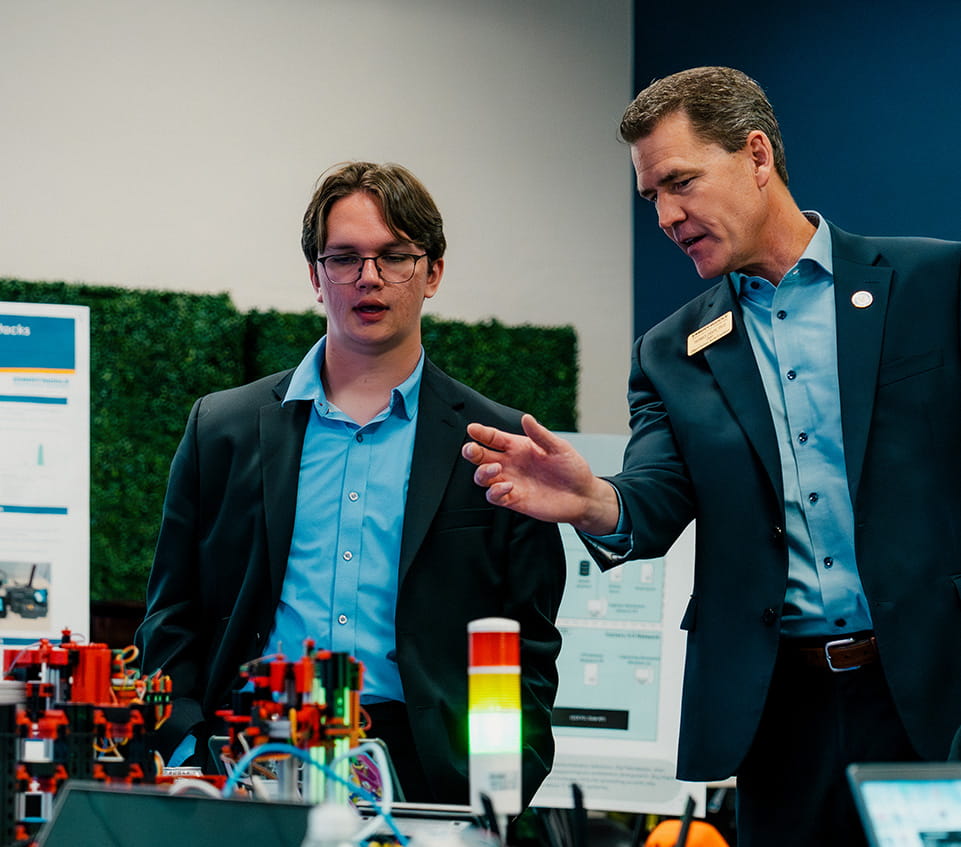 Faculty member in business attire demonstrates a small robotics and automation setup with wiring, sensors, and a stack light indicator at a display table.
