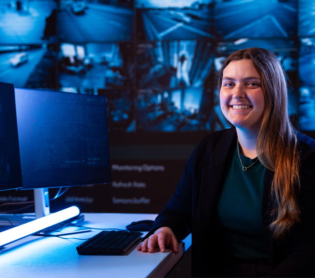 A smiling woman sits at a desk in front of a computer monitor. Behind her, a large screen displays multiple surveillance or control room images, suggesting a technology or operations setting.