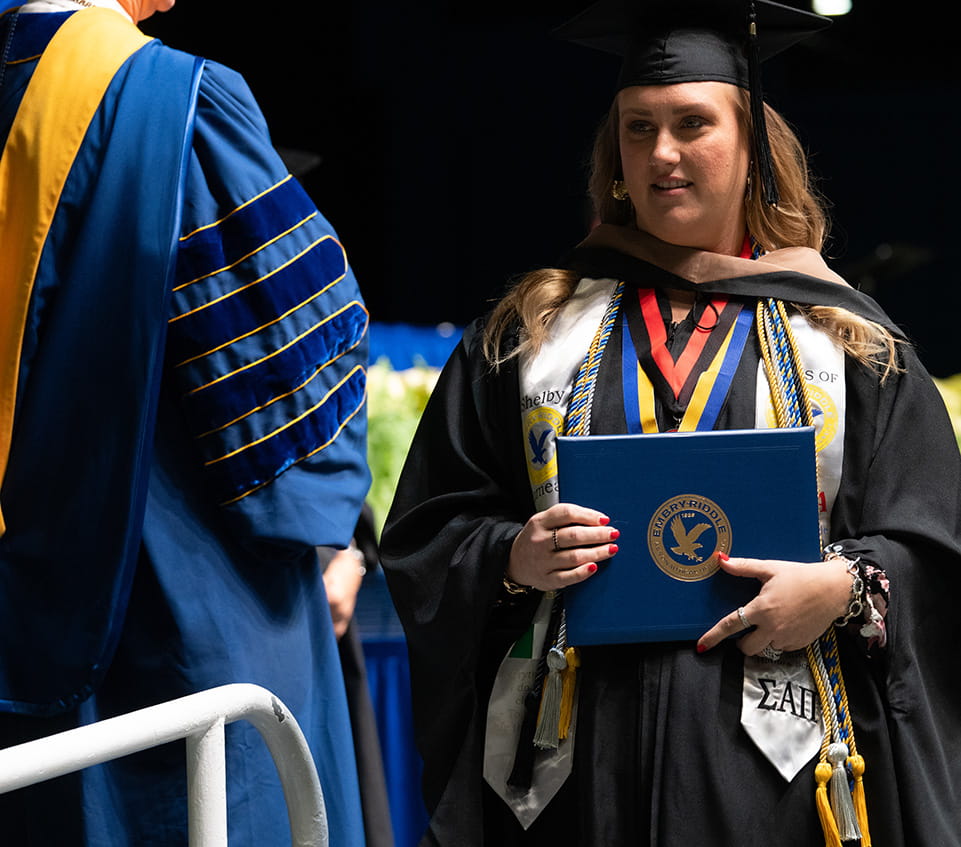 Master's graduate in cap and gown holding an Embry-Riddle diploma cover onstage, wearing honor cords, medals, and a white stole during commencement.