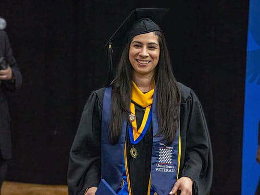 Graduate in cap and gown wearing honor cords, medals, and a United States veteran stole stands on stage smiling during commencement.
