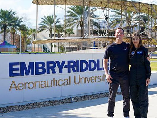Two people in matching black uniforms stand in front of Embry-Riddle Aeronautical University sign with palm trees and aircraft hangar visible behind them.