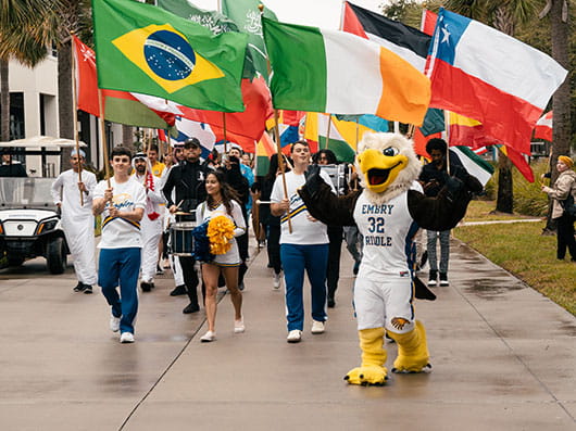 A group of people holding international flags whilst walking with ERNIE, the school mascot.
