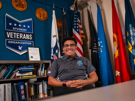 A smiling person sits behind a desk in an office decorated with military flags. A sign on the wall reads “Student Veterans Organization,” and a bookshelf filled with books is visible to the left.