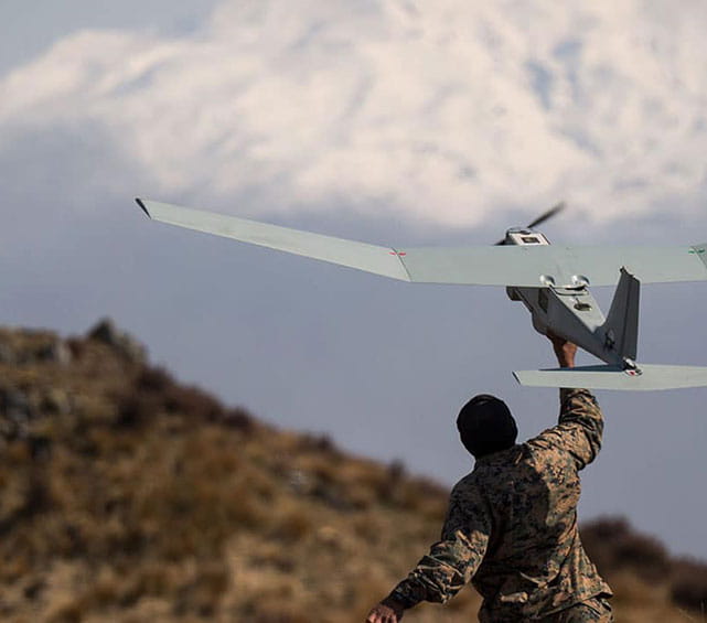 An operator in camouflage releases a hand-launched fixed-wing unmanned aerial vehicle with pusher propeller and twin-boom tail, on a rocky hillside with mountains in the background.