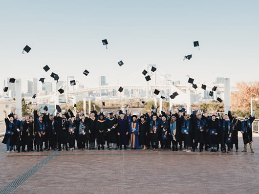 A large group of college graduates wearing graduation regalia outdoors, celebrating as they toss their caps into the air.