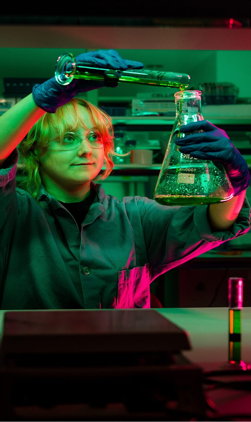 Scientist wearing gloves pours liquid from a glass beaker into an Erlenmeyer flask, holding it up at eye level to observe the reaction, with lab equipment and test tubes on a workbench in a laboratory.