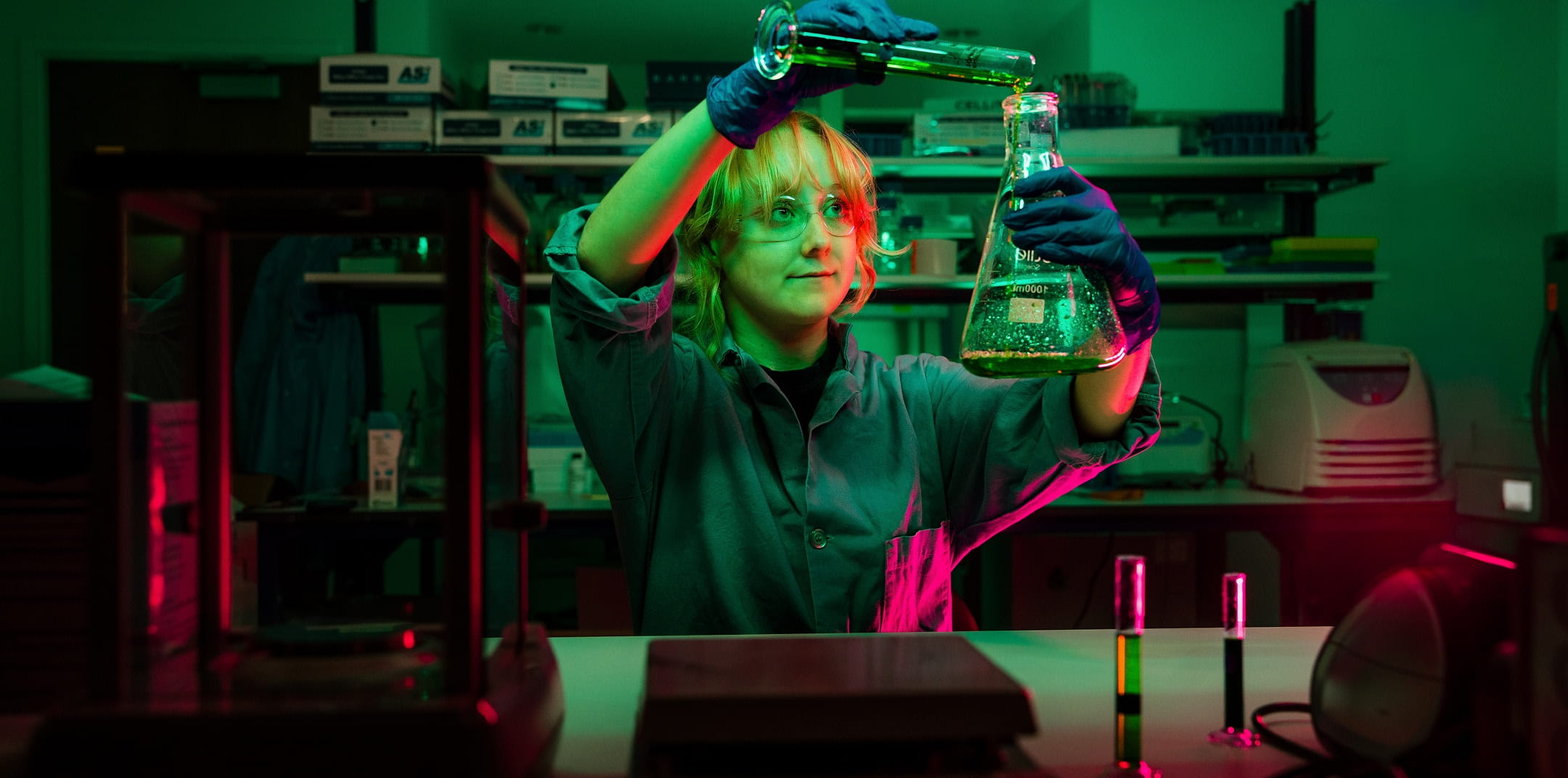 Scientist wearing gloves pours liquid from a glass beaker into an Erlenmeyer flask, holding it up at eye level to observe the reaction, with lab equipment and test tubes on a workbench in a laboratory.