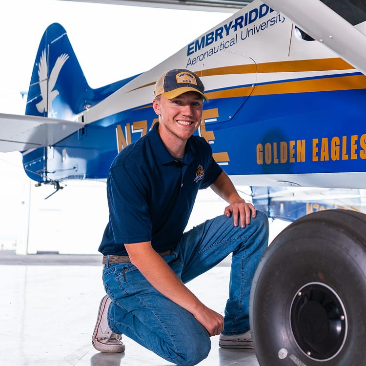A smiling man in a blue uniform kneeling next to a large commercial aircraft