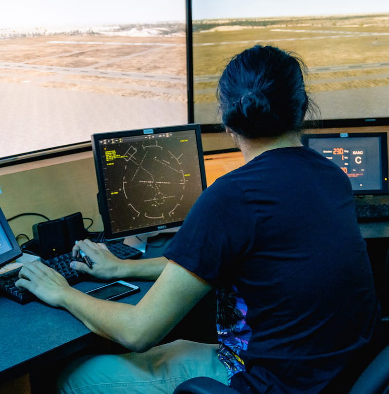 Person seated at a workstation using multiple monitors to track flight data and air traffic movements in a control room.