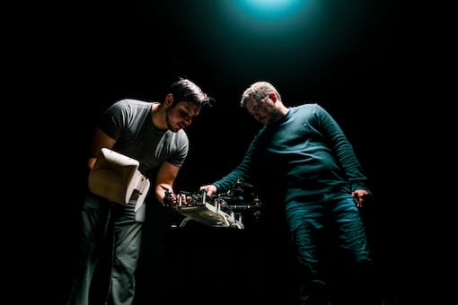 Two men examine a robotic hand under a spotlight, with one holding the device while the other looks on.