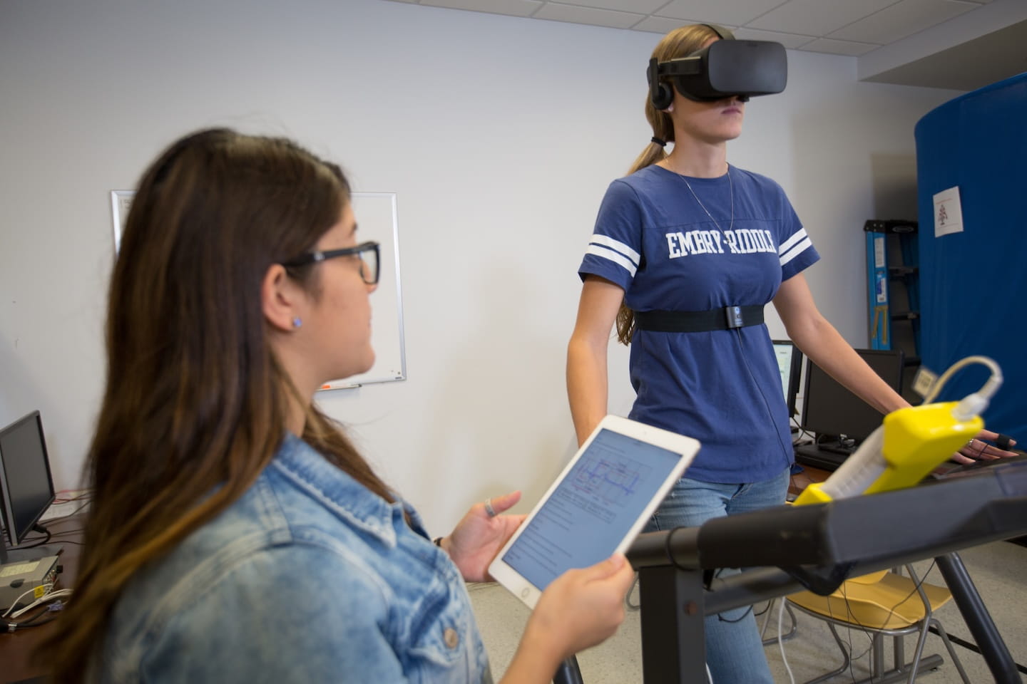 A person uses VR equipment on a treadmill while another monitors their activity nearby.