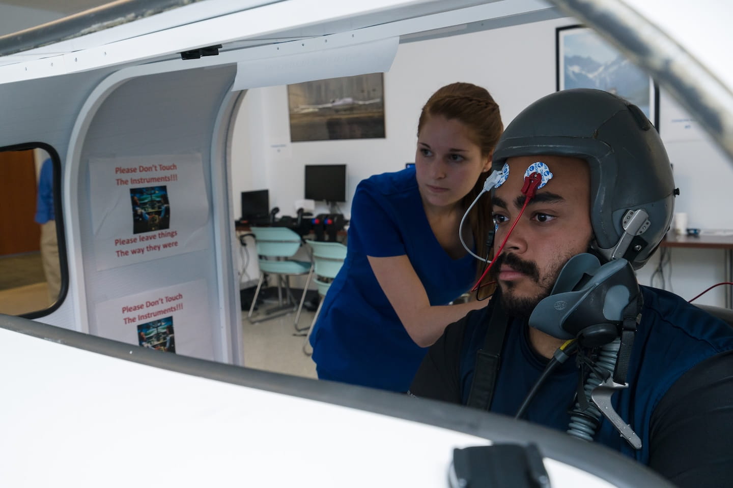 A person wearing a flight or space simulation helmet sits inside a simulator while another person stands nearby assisting them.