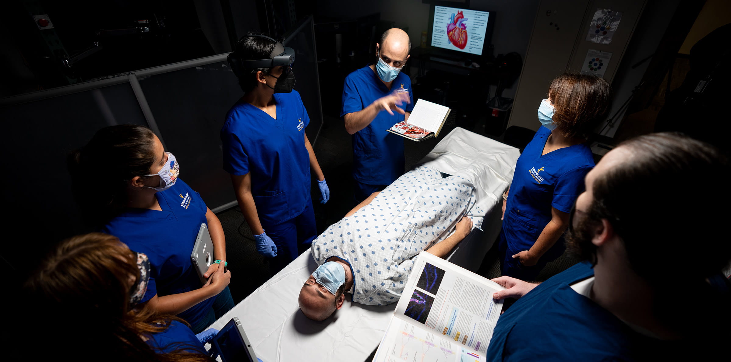 A group of clinicians stands around a hospital bed with a simulation mannequin lying on it.