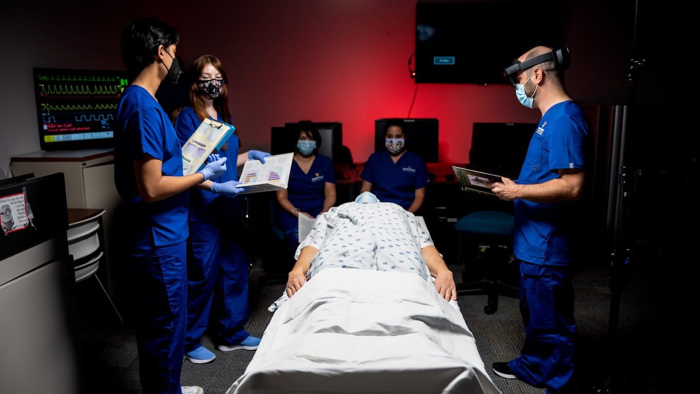Clinicians in scrubs stand around a hospital bed, monitoring a patient lying down.