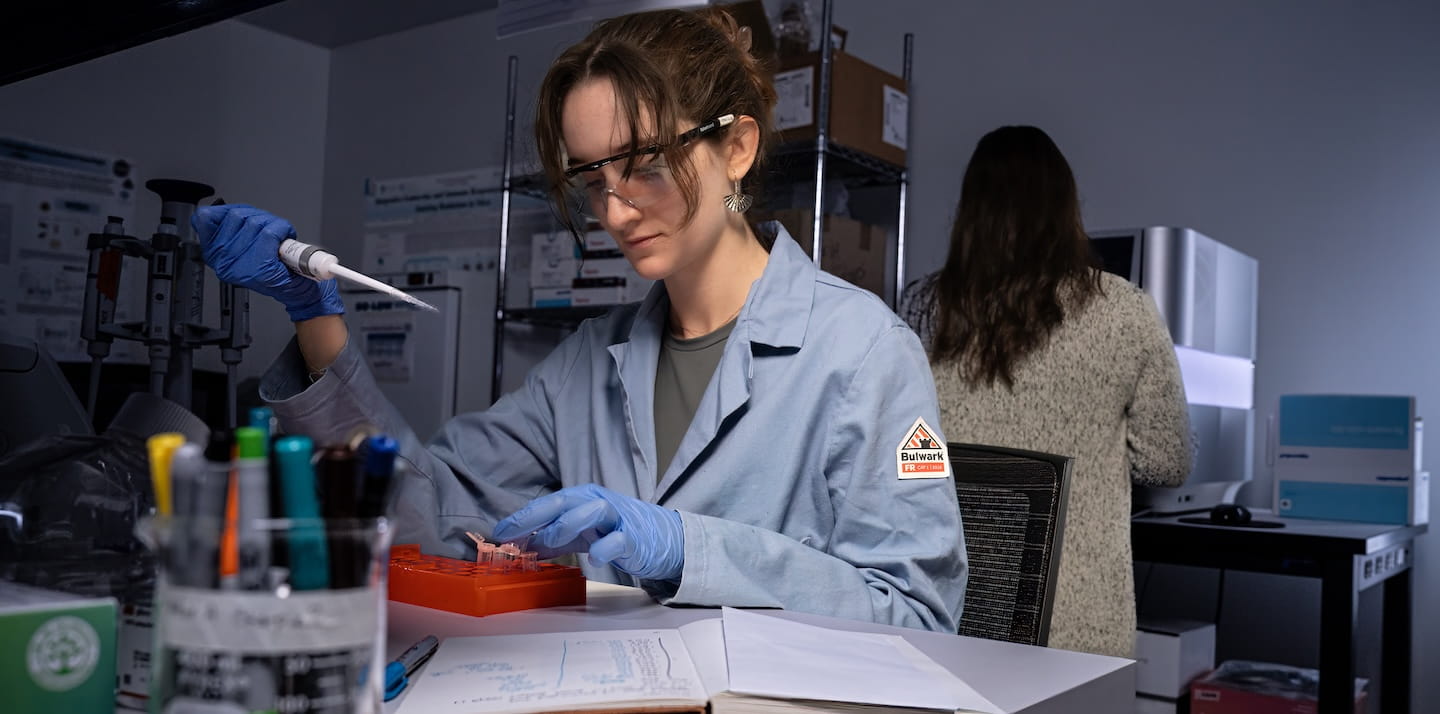 A lab worker wearing safety goggles and gloves handles equipment at a workstation.